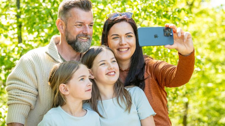 Eine Familie macht ein Selfie im Grünen. Sie lächeln glücklich in die Kamera.