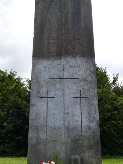 Une stèle commémorative ornée de trois croix et d'une inscription. Devant la stèle se trouvent des fleurs et une petite lanterne.