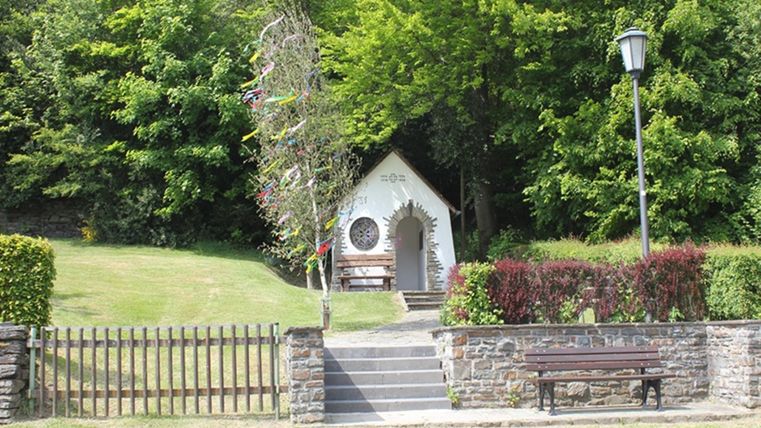 Ein kleines, charmantes Kirchlein steht umgeben von grünen Bäumen. Auf der Wiese davor lädt eine Bank zum Verweilen ein.