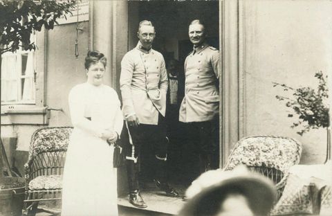 Drei Personen stehen vor einem Eingang: zwei Männer in historischen Uniformen und eine Frau in einem weißen Kleid. Im Hintergrund sieht man einige Sitzmöbel und eine blühende Pflanze.