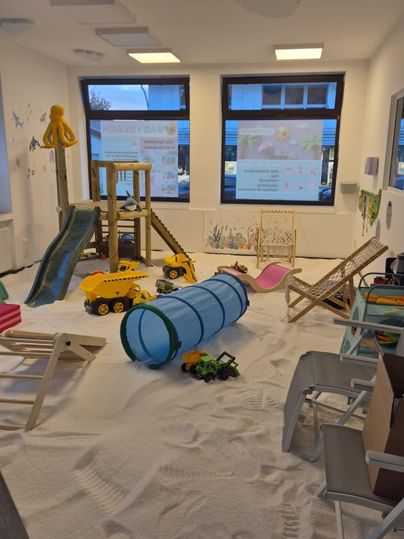 Ein Spielzimmer mit Sandboden, Spielzeugautos und einer Rutsche. In der Mitte befindet sich ein Tunnel zum Spielen und mehrere Sitzmöglichkeiten.