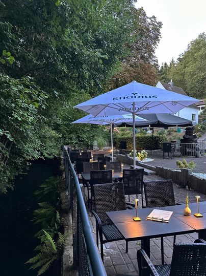 Ein gemütliches Restaurant am Fluss mit dunklen Tischen und Stühlen. Sonnenschirme bieten Schutz vor der Sonne und die Umgebung ist grün und einladend.