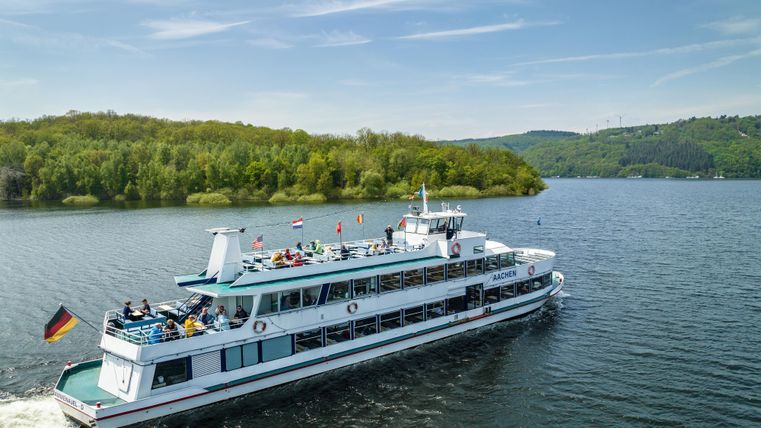 Un bateau d'excursion navigue sur un lac calme, entouré de collines verdoyantes. Il y a de nombreux passagers à bord qui profitent de la vue.