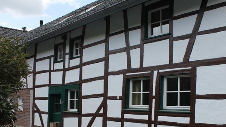 Ein traditionelles Fachwerkhaus mit weißen Wänden und braunen Balken. Die Fenster sind grün gestrichen und das Dach ist gedeckt.