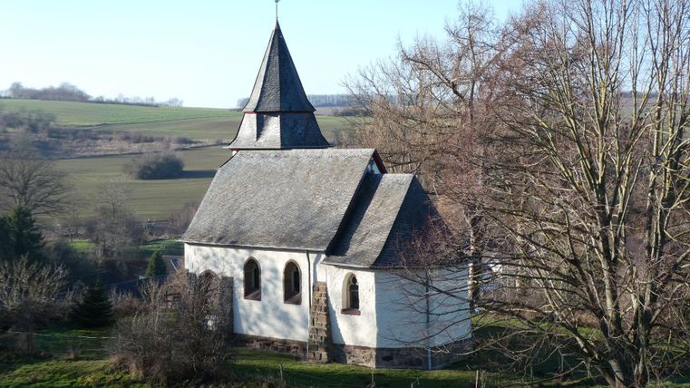 Eine kleine Kirche mit einem spitzen Turm steht in einer grünen Landschaft. Im Hintergrund sind sanfte Hügel und ein klarer Himmel zu sehen.