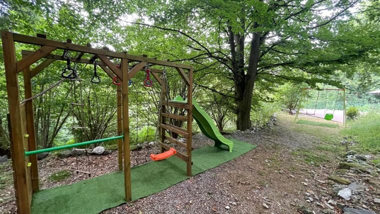 Een speelplaats van hout met een glijbaan en klimelementen is omringd door bomen. De bodem is bedekt met een groene vloerbedekking en er is een smal pad op de achtergrond.