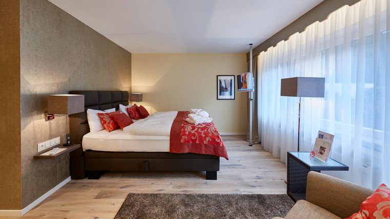 Ein modernes Hotelzimmer mit einem großen Bett und roten Akzenten. Große Fenster lassen viel Licht hinein und der Boden ist mit Holz und Teppich ausgelegt.