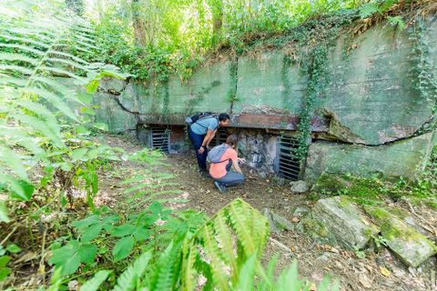 Zwei Personen erkunden einen alten Bunker, der von Pflanzen umgeben ist. Die Umgebung ist üppig und grün, was dem Bild eine geheimnisvolle Atmosphäre verleiht.