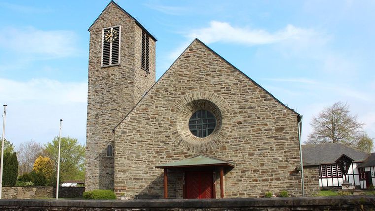 Eine steinerne Kirche mit einem hohen Turm und großen Fenstern. Umgeben von Bäumen und einer ruhigen Landschaft.