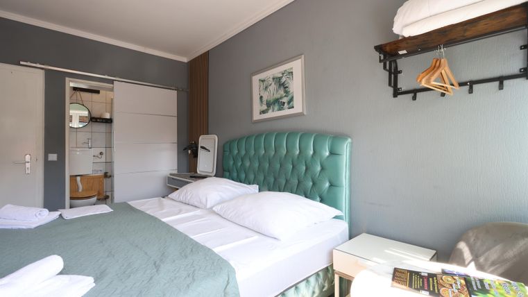 Ein modernes Zimmer mit einem grünen Bett und weißen Bettwaren. Im Hintergrund sind ein Schreibtisch und eine ausgestattete Badezimmernische zu sehen.