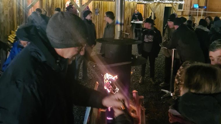 Eine festliche Szene mit Menschen, die rund um einen Holzstand stehen. Es ist dunkel, und die Umgebung ist mit Lichtern geschmückt.