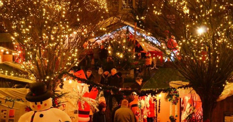 Ein festlicher Weihnachtsmarkt mit beleuchteten Ständen und einem Schneemann im Vordergrund. Viele Menschen genießen die festliche Atmosphäre.