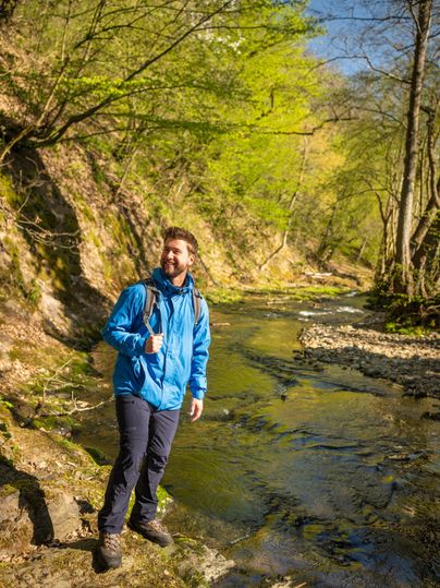 Un homme se tient au bord d'un ruisseau clair dans une forêt verte. C'est une journée ensoleillée et l'environnement semble accueillant et amical.