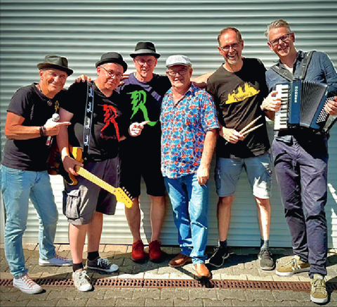 Zes muzikanten staan glimlachend voor een heldere, geribbelde muur. Ze houden muziekinstrumenten vast en dragen verschillende, kleurrijke kleding.