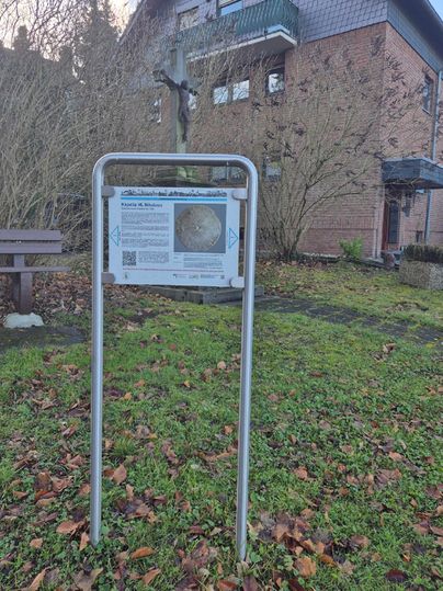 An information sign stands in a garden with autumn leaves. In the background, a building and a cross are visible.