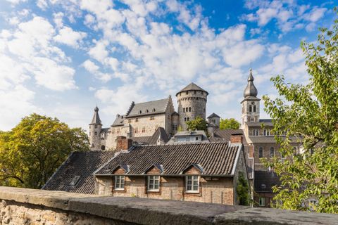 Eine beeindruckende Burg mit Türmen und Zinnen, umgeben von Bäumen. Der Himmel ist blau mit einigen Wolken.