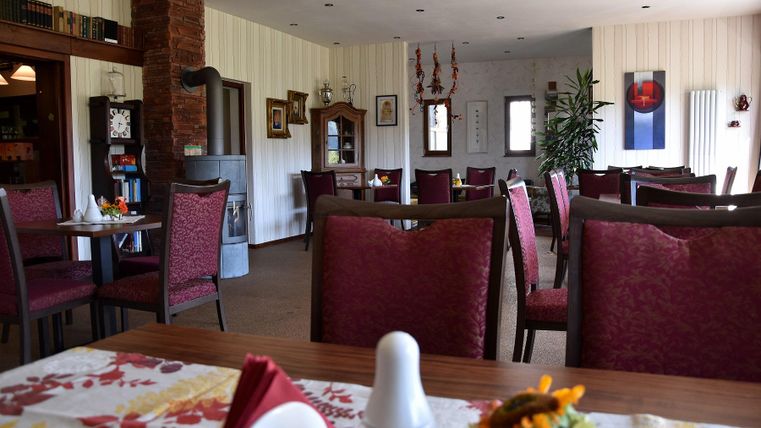 Ein gemütlicher Speiseraum mit rot gepolsterten Stühlen und runden Tischen. An den Wänden hängen Bilder und es stehen Pflanzen zur Dekoration.