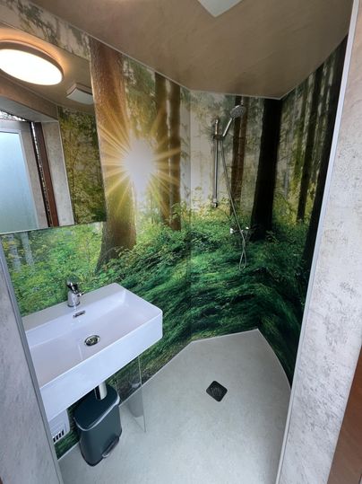 Eine moderne Dusche mit einem Waschbecken und einem Waldbild an der Wand. Das Licht scheint durch die Bäume und schafft eine beruhigende Atmosphäre.