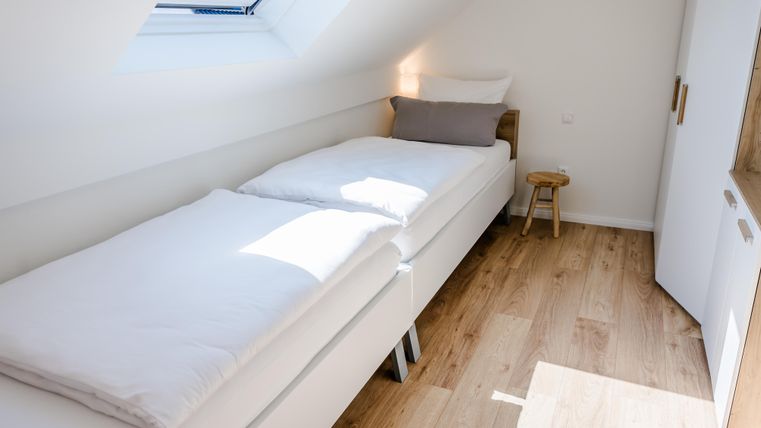Ein helles Schlafzimmer mit zwei Bettgestellen und einem kleinen Hocker. Die Wände sind weiß und der Boden ist aus Holz.