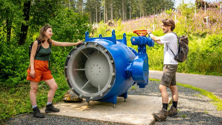Twee personen staan naast een groot blauw ventiel in de natuur. Op de achtergrond zijn bomen en bloeiende planten te zien.