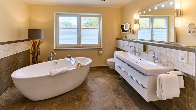 Ein modernes Badezimmer mit einer freistehenden Badewanne und einem Doppelwaschtisch. Große Fenster bieten einen Blick auf die natürliche Umgebung.
