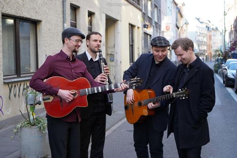 Eine Gruppe von vier Musikern spielt in einer Straße. Sie haben Gitarre und Klarinette dabei und wirken entspannt und freundlich.