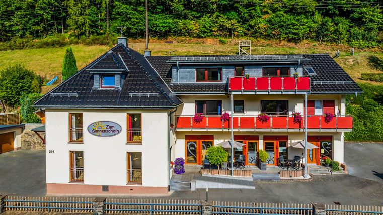 Ein charmantes Hotel mit roten Balkonen in einer grünen Umgebung. Die Fassade wirkt einladend und freundlich.