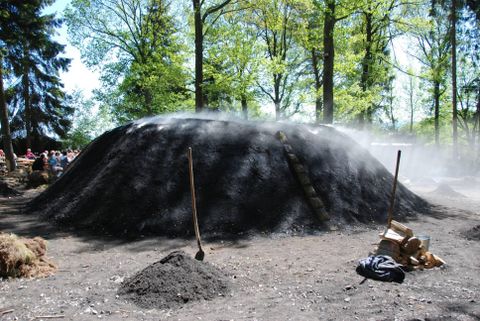 Een houtskooloven in een bosgebied, omringd door bomen. De oven is bedekt met hout en aarde, terwijl er damp opstijgt.