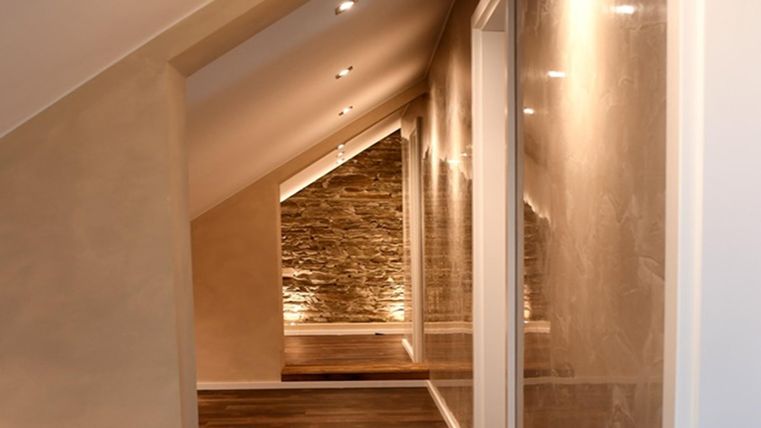 Un couloir moderne avec des murs inclinés et un éclairage agréable. Le sol est en bois et il y a un mur en pierre à l'arrière-plan.