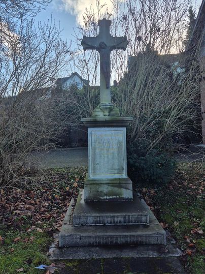 A stone cross on a pedestal, surrounded by dry bushes and faded leaves. The sky is partly cloudy.