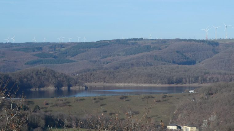 Eine ruhige Landschaft mit einem See und sanften Hügeln im Hintergrund. Am Horizont sind Windräder zu sehen.
