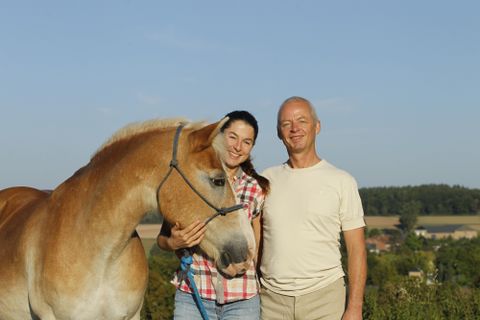 Une femme et un homme se tiennent côte à côte et sourient. À côté d'eux se trouve un cheval devant un paysage pittoresque.