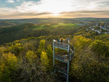 Der Krawutschketurm ist in der Mitte des Bildes und wird umrahmt von dichten Wäldern. Im Hintergrund geht die Sonne unter