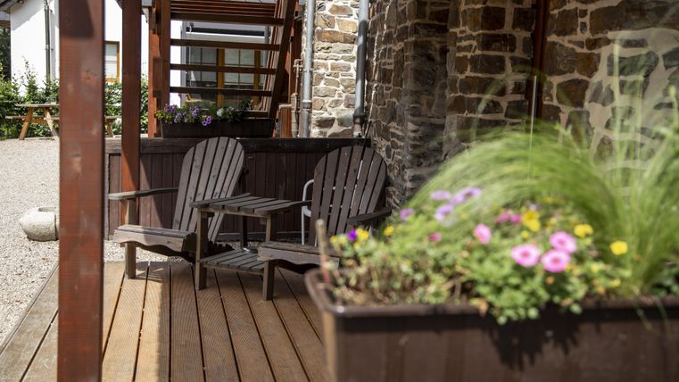 Eine gemütliche Terrasse mit zwei Stühlen und blühenden Pflanzen. Der Boden besteht aus Holz und im Hintergrund sind Steinmauern zu sehen.