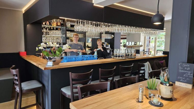 Eine moderne Bar mit Holztheke und zahlreichen Gläsern. Zwei Mitarbeiter stehen freundlich hinter der Bar.