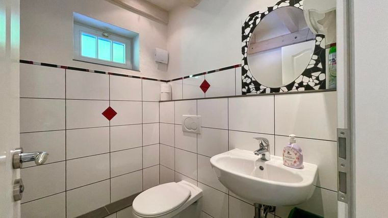 Ein kleines Badezimmer mit weißen Fliesen und einem runden Spiegel. Es gibt ein Waschbecken und ein WC, sowie eine Fensteröffnung für Tageslicht.