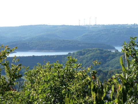 A picturesque view of a reservoir, surrounded by green hills. In the background, wind turbines can be seen.