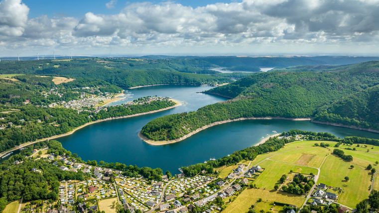 Ein weiter Blick über den Rursee mit Campingplätzen, Dörfern und Wäldern in hügeliger Umgebung.