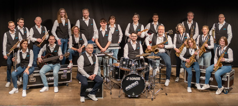 Eine große Band steht auf einer Bühne und posiert zusammen. Die Mitglieder tragen einheitliche Kleidung und halten verschiedene Musikinstrumente.