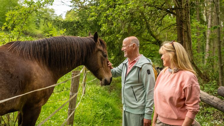 Un couple se tient sur un chemin vert et caresse un cheval. À l'arrière-plan, on peut voir des arbres et beaucoup de nature.