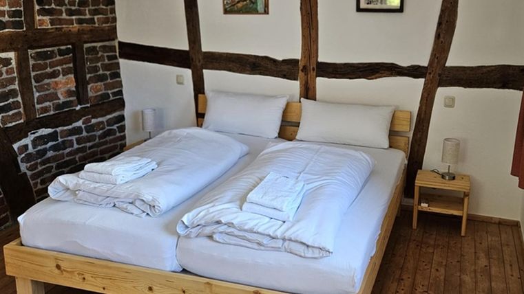 Ein gemütliches Schlafzimmer mit einem Doppelbett und frischen Bettlaken. Die Wände sind teils aus Fachwerk und die Einrichtung ist schlicht und modern.