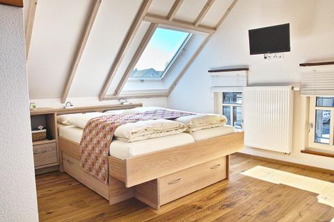 Ein gemütliches Schlafzimmer mit einem Doppelbett und Holzfußboden. Das Zimmer hat schräg verlaufende Wände, ein Fenster mit Ausblick und einen Fernseher an der Wand.