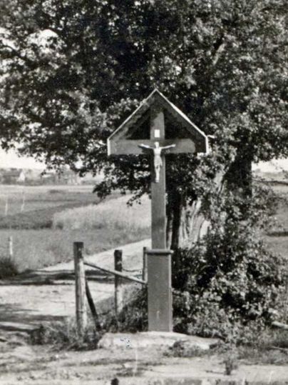 A wooden wayside cross stands along a rural path, surrounded by trees and meadows. An open landscape stretches out in the background.