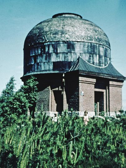 Ein historisches Observatorium mit einer gewölbten Kuppel steht vor einer grünen Landschaft. Um das Gebäude herum wachsen verschiedene Pflanzen.