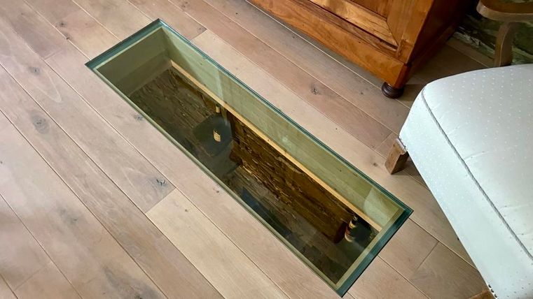 Ein rechteckiges Glasfenster im Boden zeigt eine darunterliegende Holzstruktur. Der Raum hat einen rustikalen Stil mit einem Holzfußboden und einem bequemen Sessel.