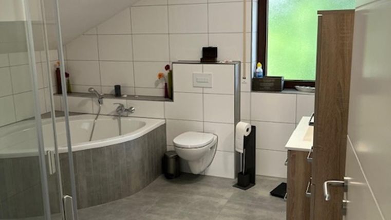 Ein modernes Badezimmer mit einer Badewanne und einer Dusche. Der Raum hat helle Fliesen und große Fenster, die Tageslicht hereinlassen.