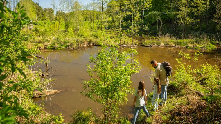 A family stands by a tranquil body of water surrounded by lush greenery. The sun is shining and it is a beautiful day in nature.