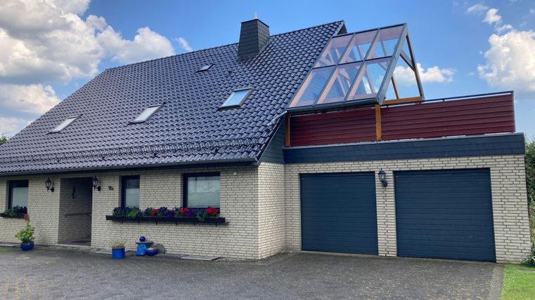 Ein modernes Haus mit einem grauen Dach und einer charakteristischen Glasgaube. Im Vordergrund sind Blumenkästen und eine Auffahrt zu sehen.
