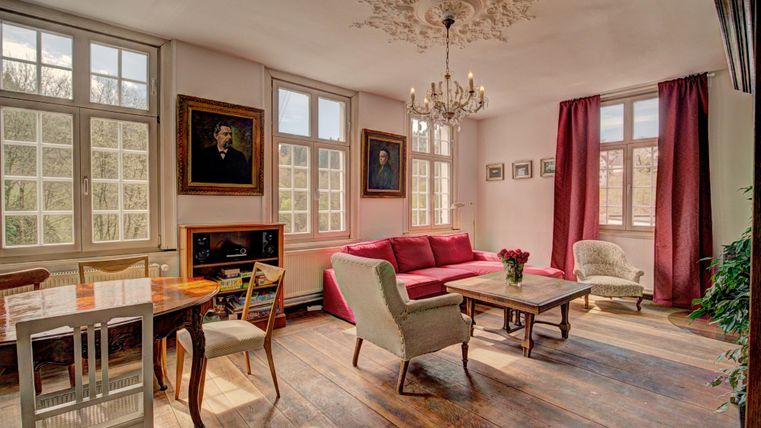 A cozy living room with large windows and a beautiful wooden floor. There is a red sofa, a dining table, and decorative pictures on the walls.