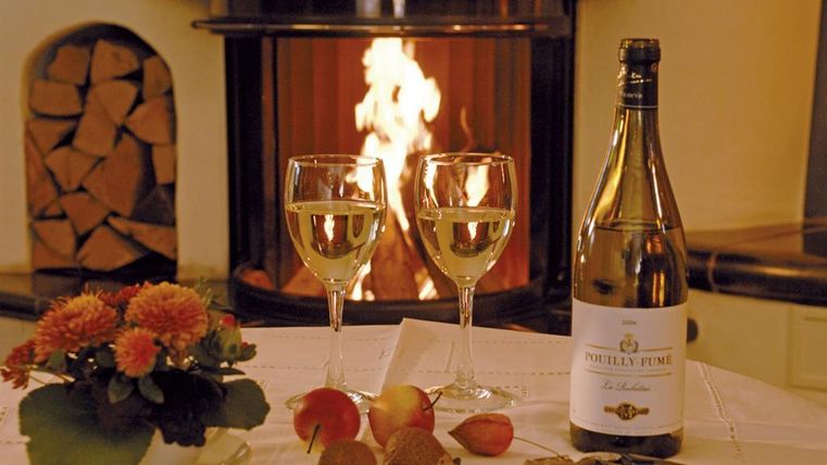 Ein gemütlicher Tisch mit zwei Weingläsern, einer Flasche Sekt und frischen Äpfeln. Im Hintergrund brennt ein Kamin und sorgt für eine warme Atmosphäre.